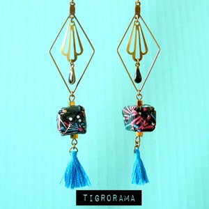 Puede incluir: Pendientes geométricos dorados con abalorios en forma de cubo con estampado negro y rosa y borlas azules.