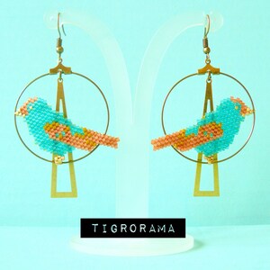 May include: A pair of turquoise and orange beaded bird earrings hanging from gold hoops. The earrings are on a white stand with the word "TIGRORAMA" printed on a black label.