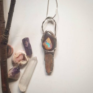 May include: A handcrafted silver pendant necklace featuring a large, iridescent opal stone set in a decorative frame. The pendant has a long, rectangular crystal below the opal. Additional gemstones and a branch are in the background.