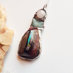 May include: A handcrafted pendant necklace featuring a large, dark brown opal with iridescent flashes of colour. The pendant also includes a light green stone and a round, pale pink stone, all set in a silver-coloured metal.
