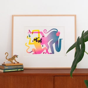 Staged photo of the "monkeys with pear" print. It's inside a light-wood frame, with a tropical looking plant in a corner, some books and a little statue of a tiger on the other.