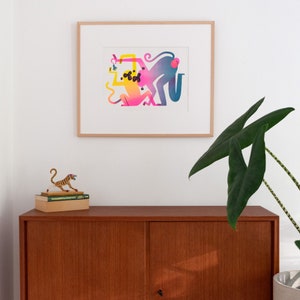 Staged photo of the "monkeys with pear" print. It's hanging on the wall, inside a light-wood frame, with a tropical looking plant in a corner, some books and a little statue of a tiger on the other.