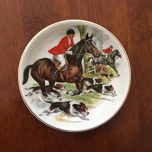 Op de afbeelding: Een witte decoratieve schotel met een gouden rand, met een jachtsc&egrave;ne. De illustratie toont een ruiter in een rode jas op een bruin paard, gevolgd door honden en andere ruiters. De schotel staat op een donkere houten ondergrond.