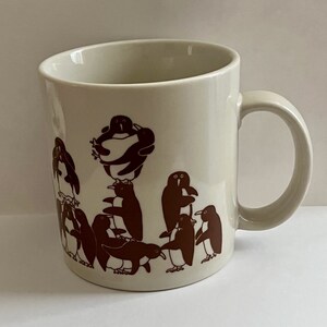 May include: Cream-colored ceramic mug with a brown penguin design. The penguins are depicted in various poses, some standing and others stacked on top of each other. The mug has a curved handle and a simple, clean design.