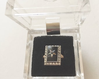May include: A black onyx ring with a five-pointed star set with blue stones, surrounded by a border of small white stones. The ring is in a clear plastic ring box with a black velvet interior.