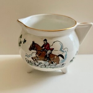 May include: White ceramic creamer with a gold rim and a depiction of a person riding a horse over a stone wall. The creamer has three legs and is decorated with shamrocks.