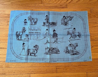 May include: A blue tea towel with a black and white illustration of a horse riding school. The illustration includes horses, riders, and a sign that reads "Thewell's Riding School".