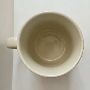 May include: Cream-colored ceramic mug with a rounded body and a small, curved handle. The interior of the mug is a slightly darker shade of cream. The mug is empty and sits on a white surface.