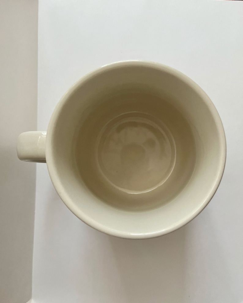 May include: Cream-colored ceramic mug with a rounded body and a small, curved handle. The interior of the mug is a slightly darker shade of cream. The mug is empty and sits on a white surface.