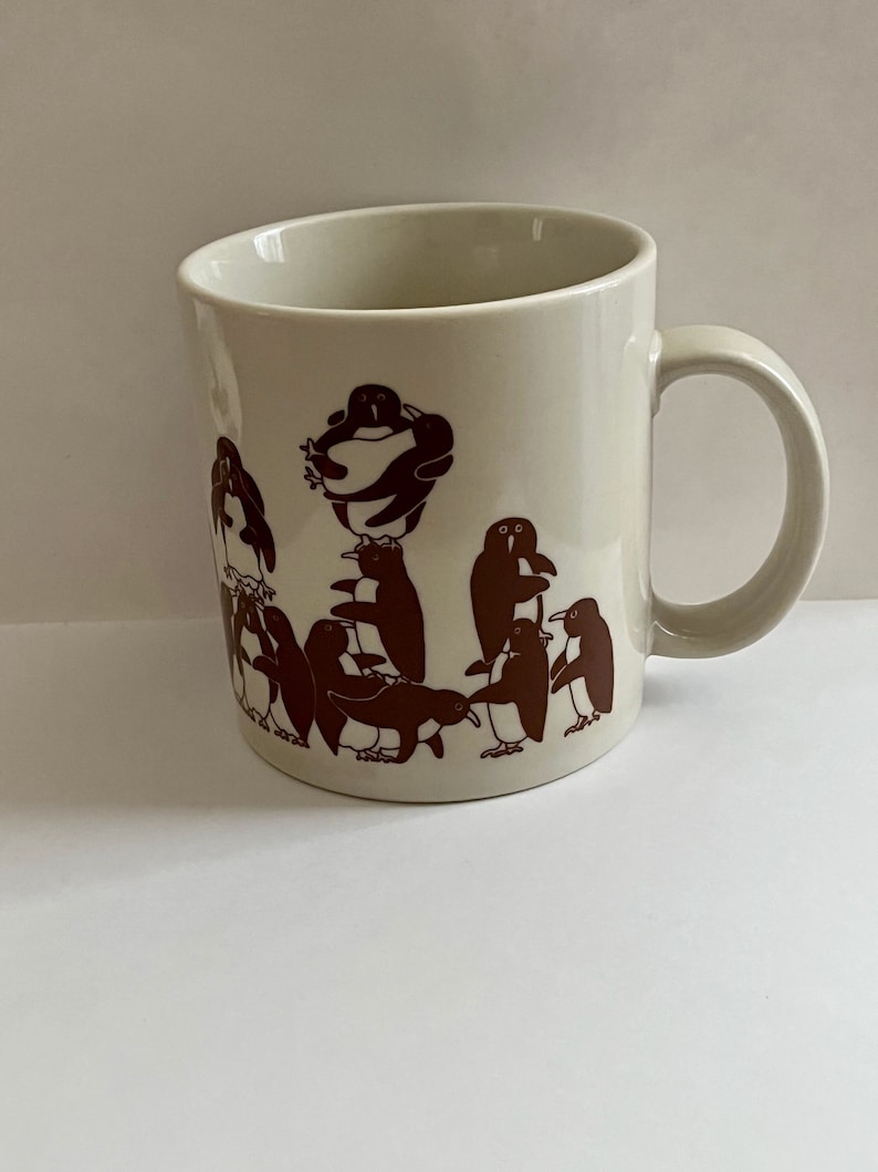 May include: Cream-colored ceramic mug with a brown penguin design. The penguins are depicted in various poses, some standing and others stacked on top of each other. The mug has a curved handle and a simple, clean design.