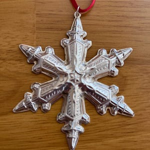 May include: A silver snowflake ornament with intricate detailing, suspended by a red ribbon. The ornament has six points, each with a textured design. A festive decoration for the holiday season.