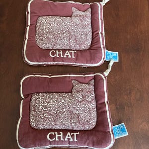 May include: Two burgundy cat-shaped pot holders with white trim and a white cat design. The word "CHAT" is printed in white on each pot holder. Each pot holder has a white loop for hanging.