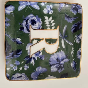 May include: Square ceramic trinket dish with a green background and a floral pattern in blue and white. The dish features a white letter "R" with gold trim and a gold border.