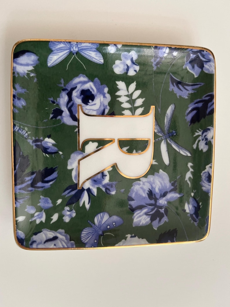 May include: Square ceramic trinket dish with a green background and a floral pattern in blue and white. The dish features a white letter "R" with gold trim and a gold border.
