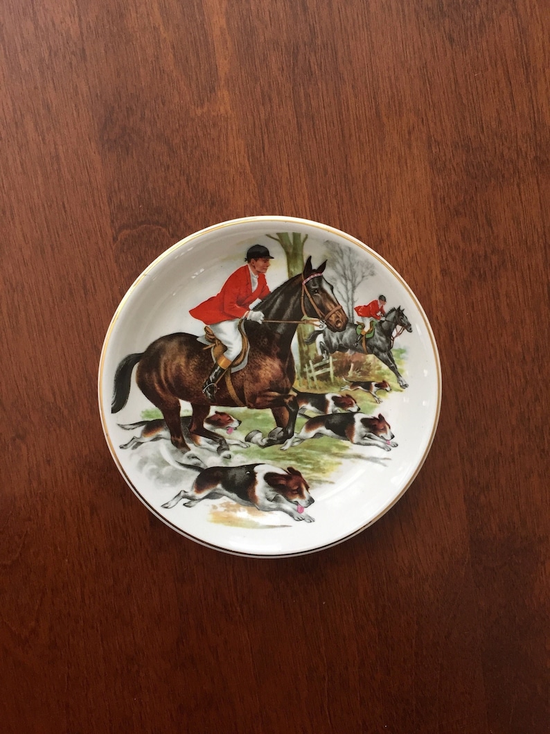 Op de afbeelding: Een witte decoratieve schotel met een gouden rand, met een jachtsc&egrave;ne. De illustratie toont een ruiter in een rode jas op een bruin paard, gevolgd door honden en andere ruiters. De schotel staat op een donkere houten ondergrond.