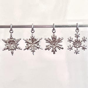 May include: Four silver snowflake charms hanging from a silver bar. Each charm has a unique snowflake design, with intricate details and varying shapes. The charms are suspended against a plain white background.
