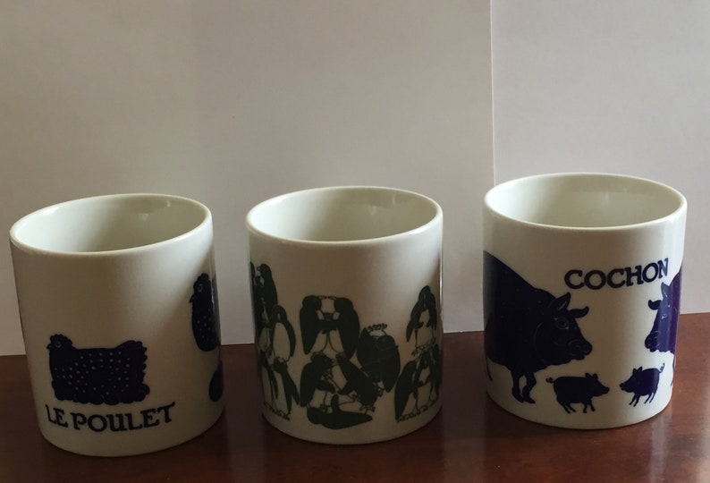May include: Three white ceramic mugs with blue illustrations and text. One mug features a chicken with the text "LE POULET". Another mug has a group of penguins. The third mug displays a cow, pigs, and the word "COCHON".