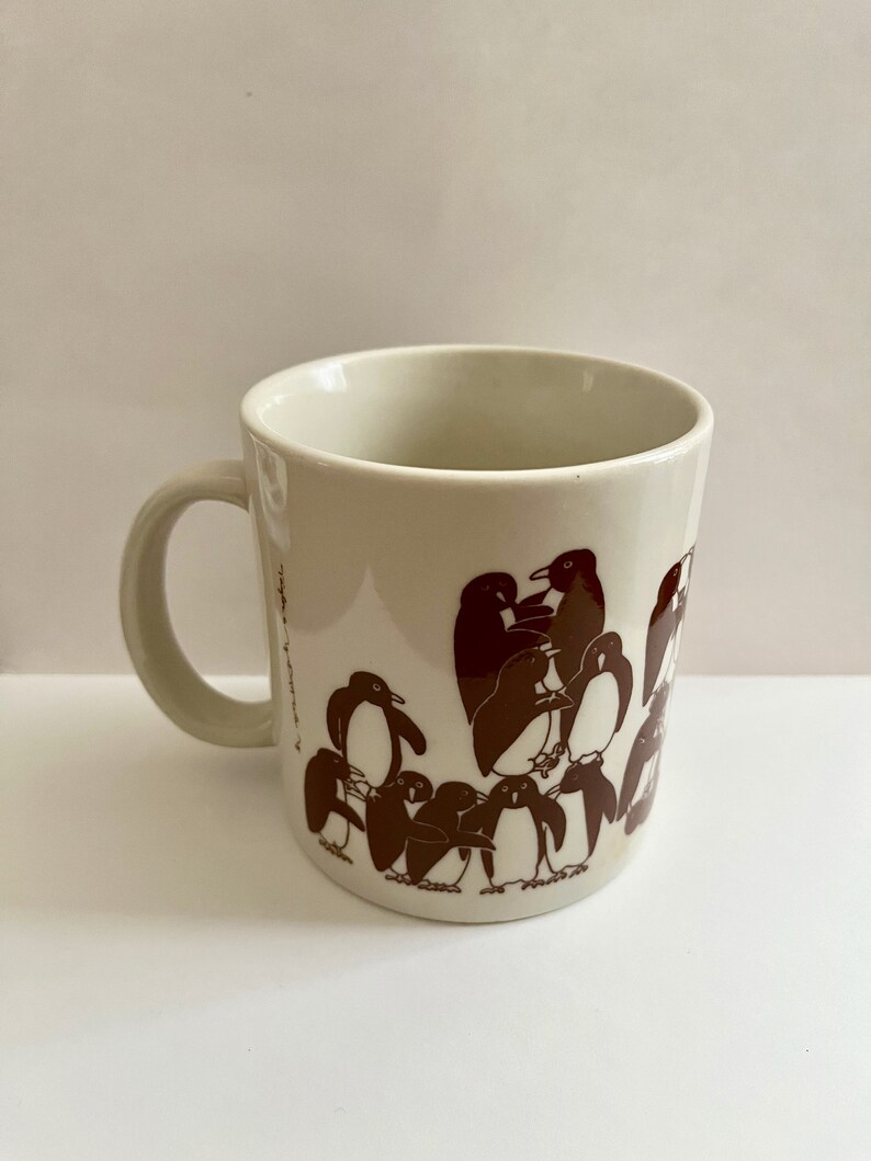 May include: Cream-colored ceramic mug with a brown penguin illustration. The mug features a curved handle and a cylindrical shape. The penguin design wraps around the mug, creating a playful and whimsical aesthetic.