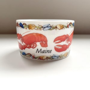 May include: A white ceramic bowl featuring a repeating pattern of red lobsters and various seashells around the top and bottom edges. The word Maine is printed in black script in the center of the bowl between the lobster illustrations.