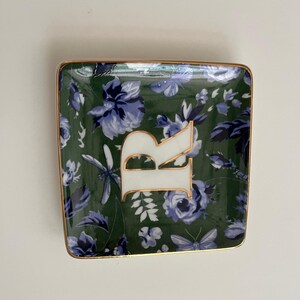 May include: Square trinket dish with a dark green floral pattern and gold trim. The dish features a large white letter "R" with gold accents. The floral design includes blue flowers, butterflies, and dragonflies.