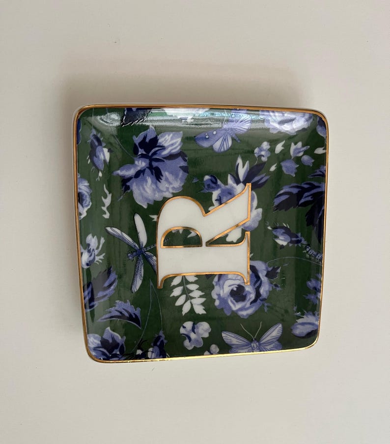 May include: Square trinket dish with a dark green floral pattern and gold trim. The dish features a large white letter "R" with gold accents. The floral design includes blue flowers, butterflies, and dragonflies.