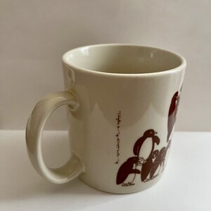 May include: Cream-colored ceramic mug featuring a brown penguin illustration. The mug has a curved handle and the text "Taylor & Yvette" printed vertically on the side. The penguin design includes several penguins in various poses.