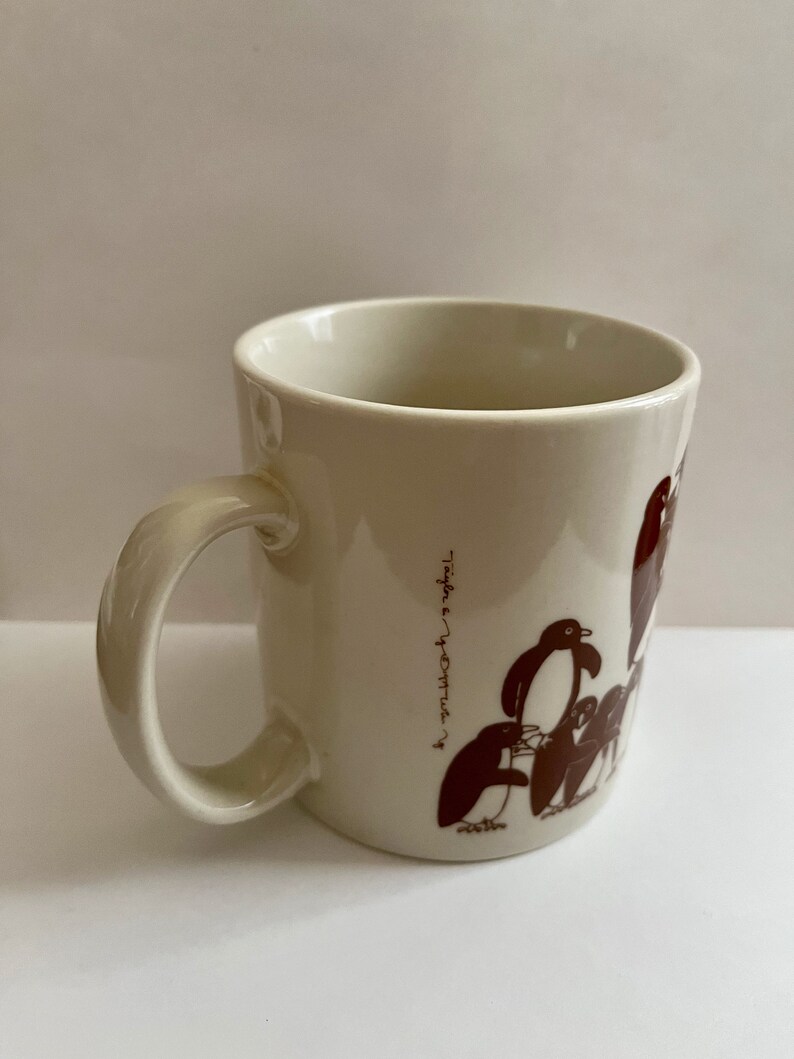 May include: Cream-colored ceramic mug featuring a brown penguin illustration. The mug has a curved handle and the text "Taylor & Yvette" printed vertically on the side. The penguin design includes several penguins in various poses.