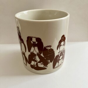 May include: A cream-colored ceramic mug featuring a repeating pattern of brown penguin illustrations. The penguins are depicted in various poses, creating a playful and whimsical design. The mug has a simple, cylindrical shape.