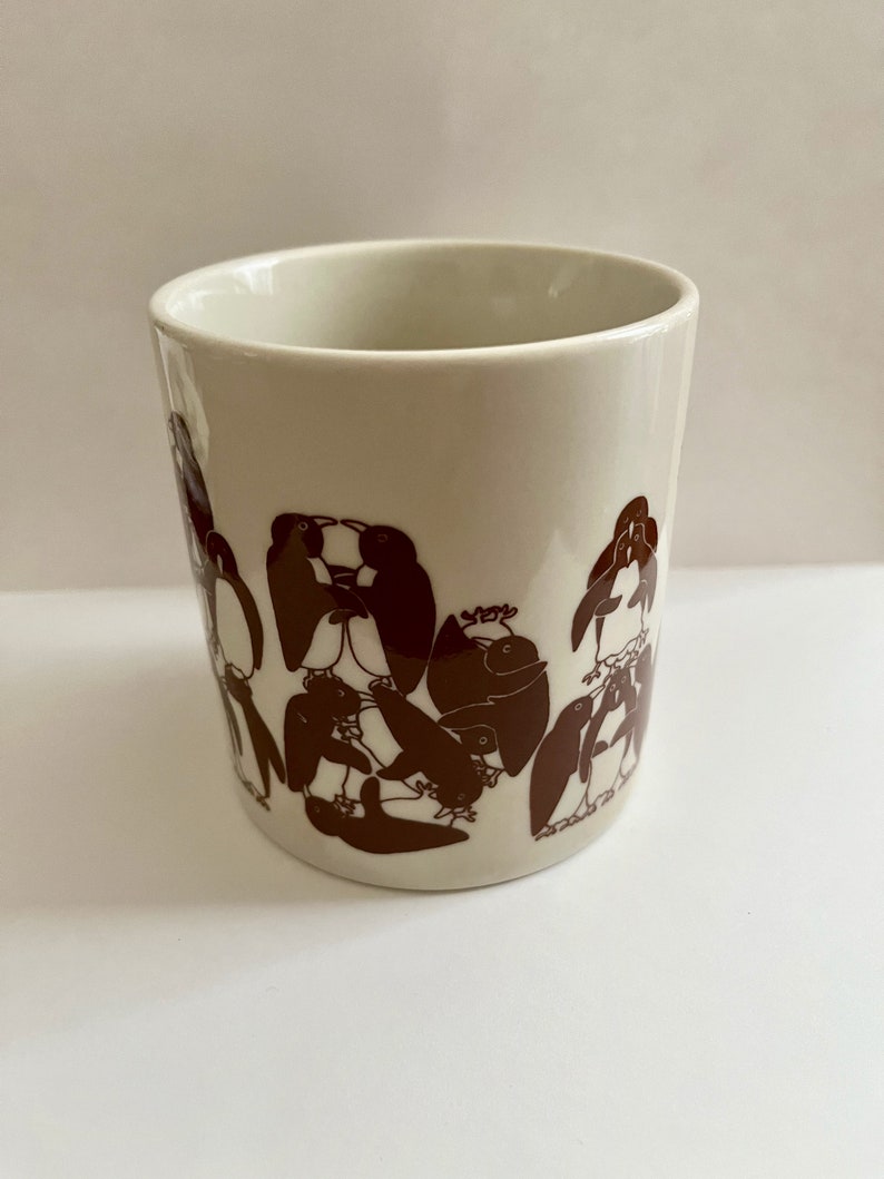 May include: A cream-colored ceramic mug featuring a repeating pattern of brown penguin illustrations. The penguins are depicted in various poses, creating a playful and whimsical design. The mug has a simple, cylindrical shape.