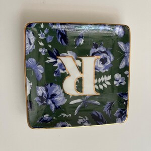 May include: Square decorative dish with a green floral pattern and a gold trim. The dish features a white letter "R" with a gold outline. The floral design includes blue flowers and butterflies.
