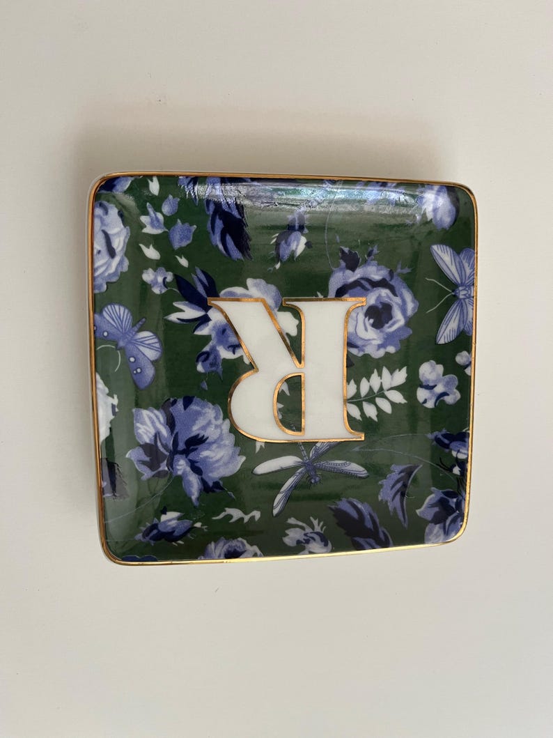 May include: Square decorative dish with a green floral pattern and a gold trim. The dish features a white letter "R" with a gold outline. The floral design includes blue flowers and butterflies.