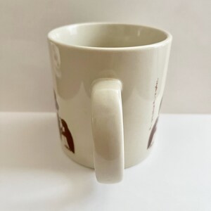 May include: Cream-colored ceramic mug with a curved handle. The mug features brown illustrations of figures and text. The mug is set against a white background.
