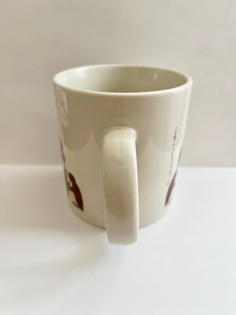May include: Cream-colored ceramic mug with a curved handle. The mug features brown illustrations of figures and text. The mug is set against a white background.