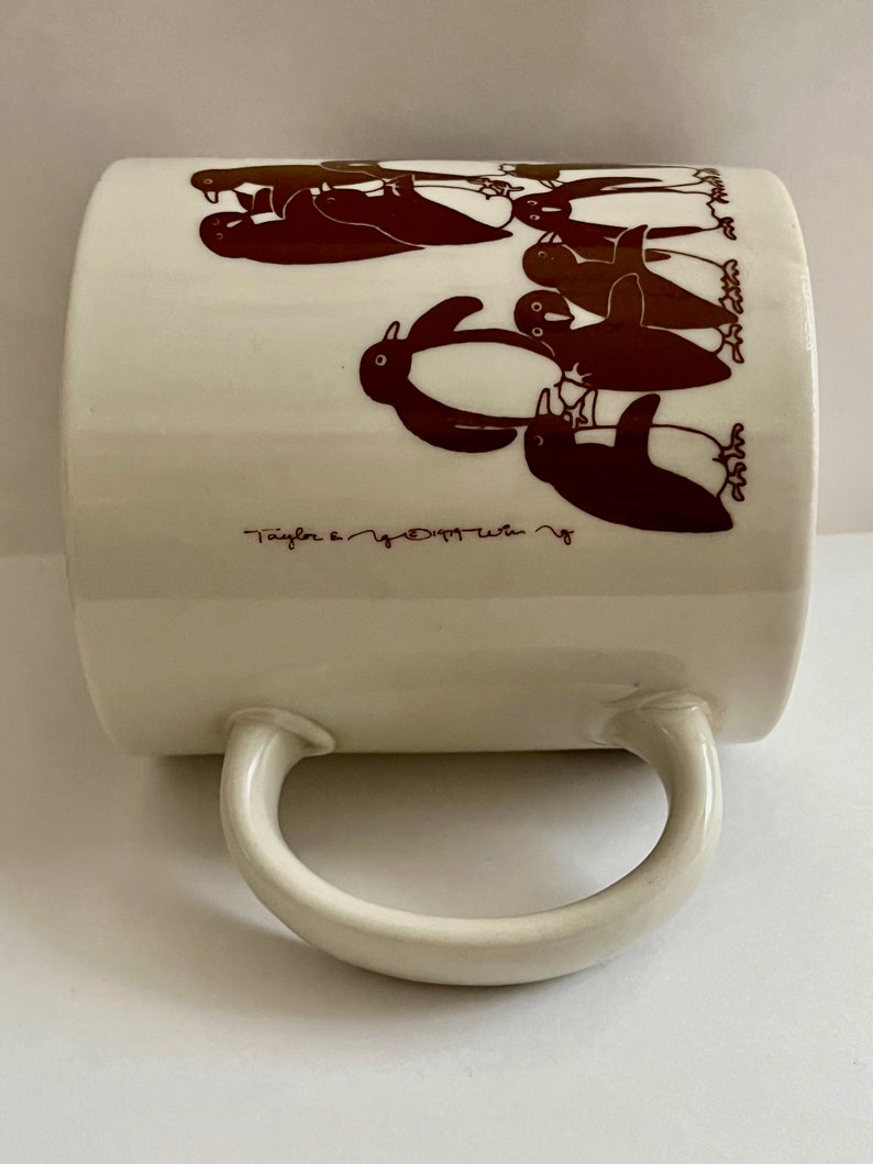 May include: Cream-colored ceramic mug featuring a repeating pattern of brown penguins. The mug has a curved handle and the text "Taylor & Co." is printed near the bottom. A charming and whimsical design for a coffee or tea cup.