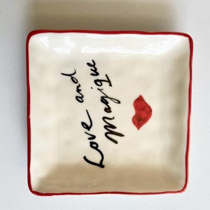 Op de afbeelding: Vierkant, cr&egrave;mekleurig keramisch schaaltje met een rode rand. Het schaaltje heeft de woorden "Love and Magique" in zwart schrift, samen met een rode lipafdruk. Een decoratief item.