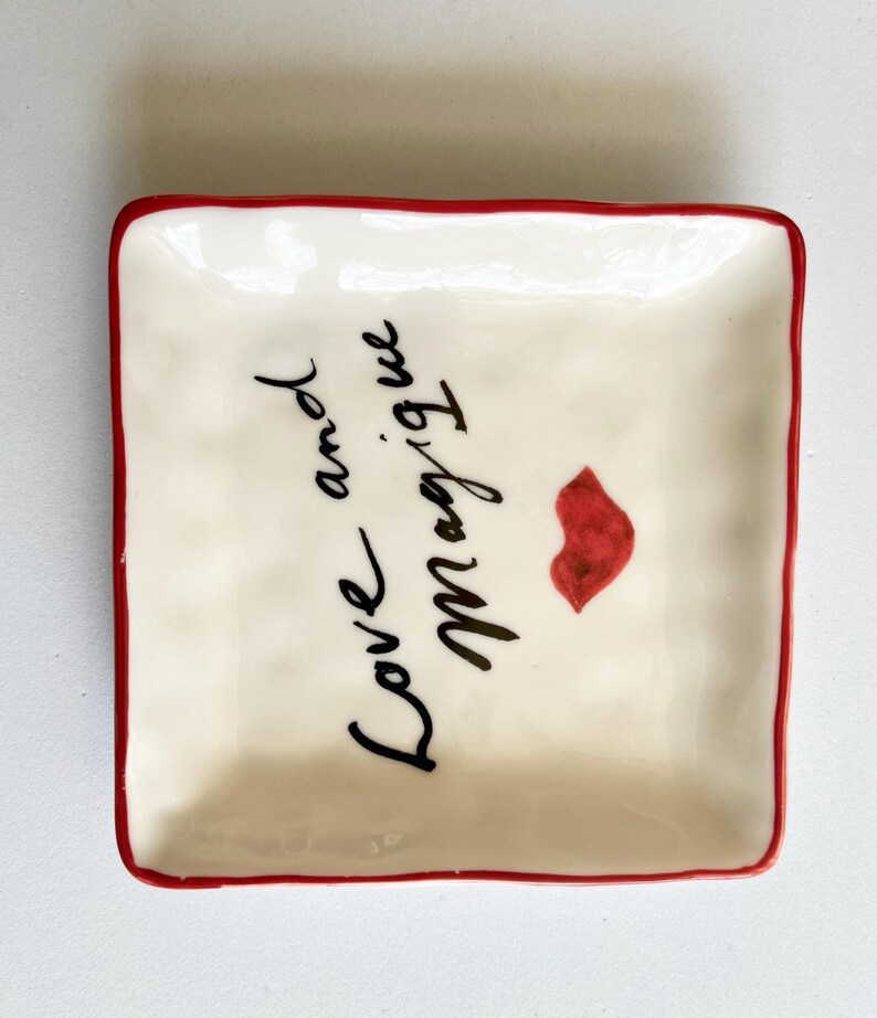Op de afbeelding: Vierkant, cr&egrave;mekleurig keramisch schaaltje met een rode rand. Het schaaltje heeft de woorden "Love and Magique" in zwart schrift, samen met een rode lipafdruk. Een decoratief item.