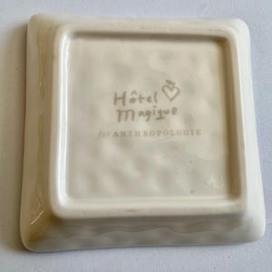 Op de afbeelding: Off-white vierkante keramische schaal met afgeronde hoeken. De schaal heeft de tekst "H&ocirc;tel Magique" en "for ANTHROPOLOGIE" in een lichtbruine kleur. Een klein hart- en bladontwerp staat boven het woord "Magique".