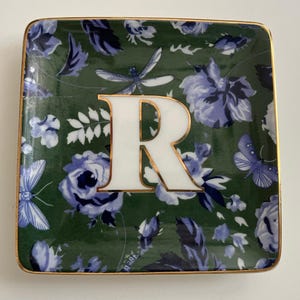 May include: Square ceramic trinket dish with a green floral pattern and a gold rim. The dish features a large white letter "R" with a gold outline, and illustrations of blue flowers, butterflies, and dragonflies.