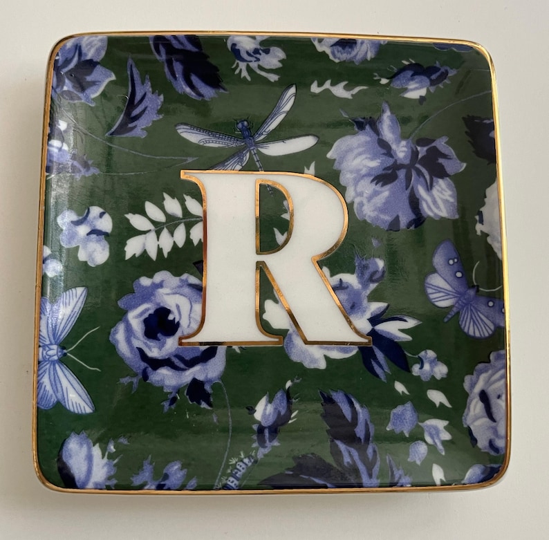 May include: Square ceramic trinket dish with a green floral pattern and a gold rim. The dish features a large white letter "R" with a gold outline, and illustrations of blue flowers, butterflies, and dragonflies.