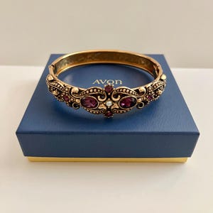 May include: A gold-toned hinged bracelet with ornate detailing and embedded purple gemstones. The bracelet is resting on a blue box with the brand name "AVON" visible. A classic piece of jewelry.