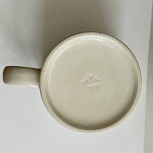 May include: Off-white ceramic mug with a rounded base and a curved handle. The bottom of the mug has a circular design with the letters "JV" and a star-like symbol. The mug is set against a plain white background.