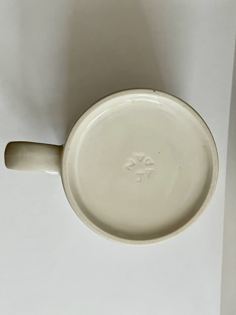 May include: Off-white ceramic mug with a rounded base and a curved handle. The bottom of the mug has a circular design with the letters "JV" and a star-like symbol. The mug is set against a plain white background.
