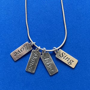 May include: Silver-toned necklace with four rectangular charms. Each charm has a word engraved: "Love", "Dance", "Live", and "Sing". The necklace is on a blue background.