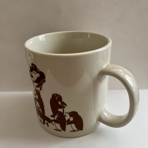 May include: Cream-colored ceramic mug featuring a brown penguin design. The mug has a rounded handle and a simple, clean design. The penguin illustration is repeated around the mug.