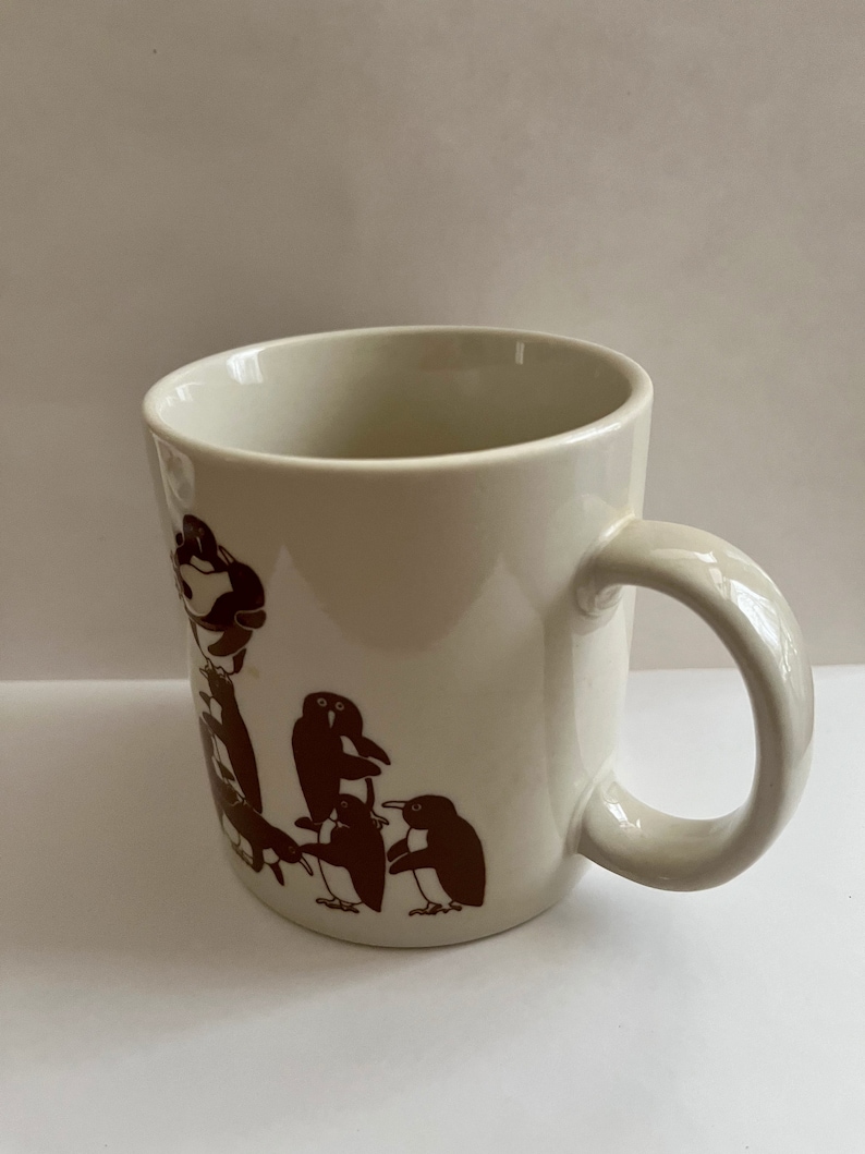 May include: Cream-colored ceramic mug featuring a brown penguin design. The mug has a rounded handle and a simple, clean design. The penguin illustration is repeated around the mug.