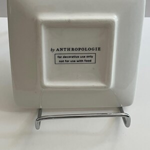 May include: A square, off-white decorative plate with rounded corners, supported by a silver metal stand. The underside of the plate features the text "by ANTHROPOLOGIE" and a warning: "for decorative use only not for use with food."