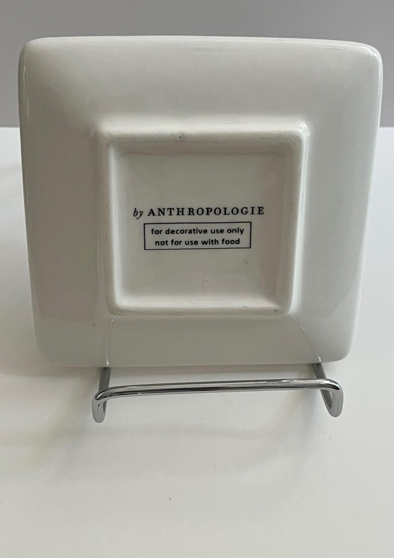 May include: A square, off-white decorative plate with rounded corners, supported by a silver metal stand. The underside of the plate features the text "by ANTHROPOLOGIE" and a warning: "for decorative use only not for use with food."