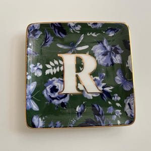 May include: Square decorative dish with a green floral pattern, featuring blue and white flowers, butterflies, and dragonflies. The dish has a gold trim and a large white letter "R" in the centre.