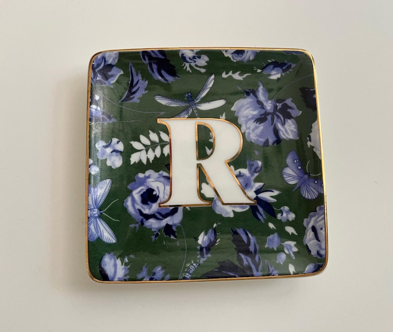 May include: Square decorative dish with a green floral pattern, featuring blue and white flowers, butterflies, and dragonflies. The dish has a gold trim and a large white letter "R" in the centre.
