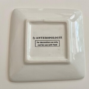 May include: A square, white Anthropologie decorative dish. The dish has rounded corners and a slightly raised square centre. Text on the bottom reads "by ANTHROPOLOGIE" and "for decorative use only not for use with food."